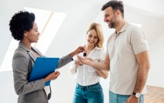 A woman in a suit hands over keys to a happy couple in a bright, wood-floored room.