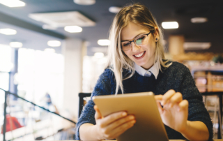 Person with glasses and light-colored hair smiling at a tablet in a cozy, modern setting