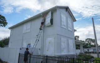 Image of two people installing protective coverings on windows of a house before a hurricane.