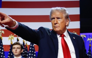 A man in a dark suit and red tie points emphatically against a backdrop of american flags.