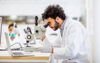 Man in a lab coat examining under a microscope with a laptop nearby