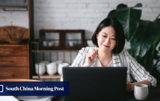 A person smiling while working on a laptop in a cozy home workspace with a large plant in the background.