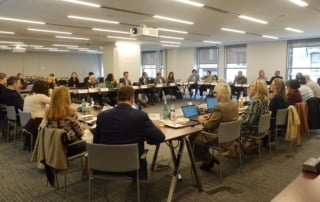 A group meeting in progress with attendees seated around tables.