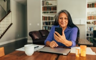 An older woman sitting at a wooden table in a cozy living room environment, engaging in a telehealth session via smartphone.