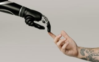 Robotic hand and tattooed human hand almost touching against neutral gray background, symbolizing human-technology collaboration in ai for cancer treatment.