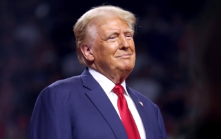 Image of a man wearing a suit and tie with a flag pin, standing indoors with a slight smile.