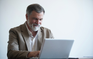 Man with gray hair and a beard wearing a beige blazer, seated at a desk using a laptop.