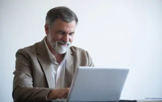 48Th Edition Of Florida Real Estate Pre-License Textbook: A Must-Have Resource 4 Man with gray hair and a beard wearing a beige blazer, seated at a desk using a laptop.