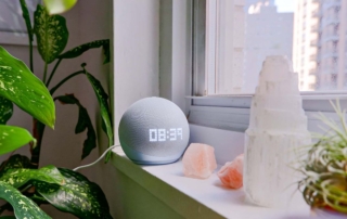 A smart speaker with a display showing the time, on a windowsill with crystals and a plant.