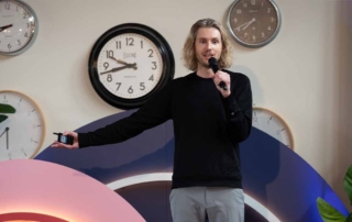 A person with long, blonde hair speaks on a stage, holding a microphone with clocks and colorful arcs in the background.