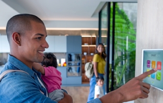 A man interacting with a smart home control panel while holding a baby, in a modern open-concept house