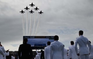 Six jet planes flying in v formation with naval personnel in the foreground