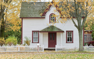 Small white house with red trim, surrounded by autumn trees and a white picket fence