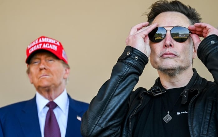 Two men in discussion: one in a leather jacket, the other in a suit with a red cap