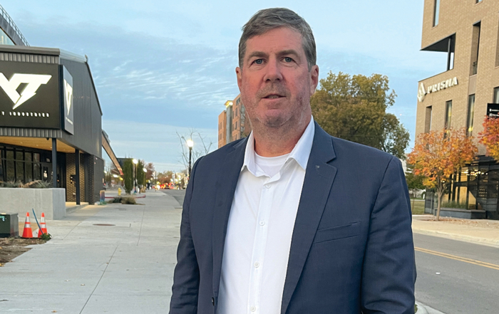 A man in a blue suit and white shirt stands on a sidewalk in an urban area with modern buildings and autumn foliage.