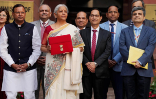 Group of people in formal attire standing outdoors, central figure in a white saree with a red briefcase