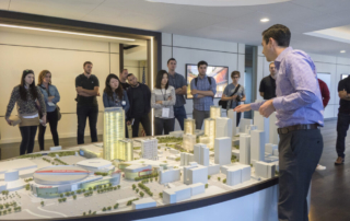 A group of people in a professional setting discuss an architectural model of a modern urban development.