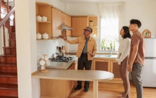 A bright, modern kitchen with three people; an older man holding a tablet explains kitchen features to a young couple.