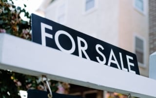 A "for sale" sign symbolizing changes in the real estate market, with a home and foliage in the background.