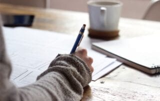 Hand writing notes on paper with a pen, surrounded by a cozy setting with a wooden table and a coffee mug