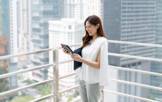 Woman on a balcony in a city, smiling and looking at a tablet, representing real estate and technology engagement.