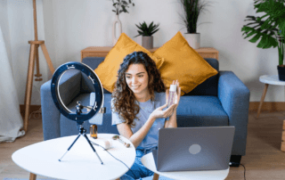 Young woman sitting on the floor in a living room, holding a cosmetic bottle with products on a table and a laptop, recording a video with a ring light.
