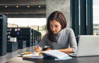 A young woman in a gray turtleneck writing in a notebook at a library desk with an open book and laptop.