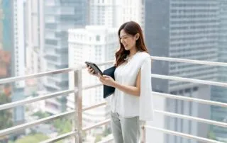 A person in a white outfit stands on a balcony, overlooking a cityscape of modern skyscrapers, interacting with a tablet.