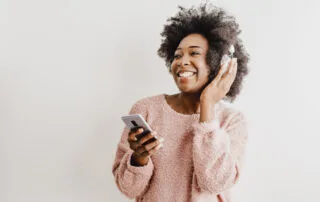 A person smiling and using a smartphone with headphones, wearing a pink sweater.
