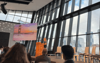 Speaker presenting 2026 emerging trends in real estate on stage at a conference with city skyline visible through large glass windows.