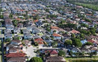 Aerial view of a large suburban neighborhood with rows of single-family homes, tree-lined streets, and backyards.
