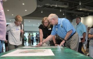 A group of adults gathers around a table, closely reviewing large printed maps or plans during an indoor community meeting.