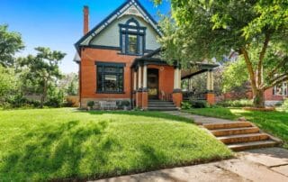 Historic red brick house with large front porch, gabled roof, and lush green lawn on a sunny day
