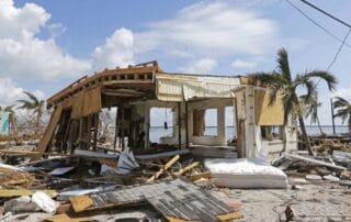 Severely damaged coastal house surrounded by storm debris and fallen palm trees after a hurricane.