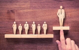 Wooden cutout figures of business people standing on blocks, with one figure elevated on a separate block being lifted by a hand, symbolizing leadership or promotion.