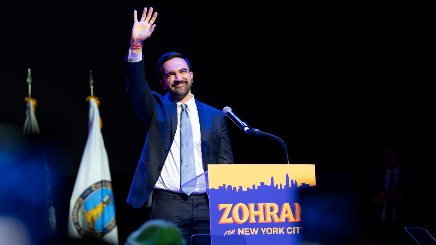Zohran Mamdani Elected As First Muslim And South Asian Mayor Of Nyc 6 Zohran mamdani waving from a podium after winning new york city mayoral race, wearing a suit with a blue tie, with a "zohran for new york city" sign in foreground