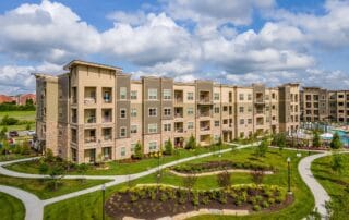 Modern apartment complex with landscaped walking paths, green lawns, and outdoor pool under a partly cloudy sky