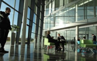 Business professionals talking and working in a modern glass office lobby with large windows and lounge seating.
