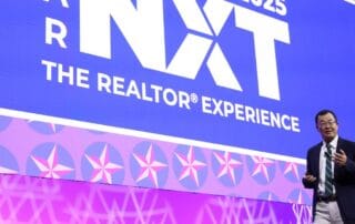 Speaker presenting on stage in front of a large “nxt – the realtor experience 2025” conference screen.