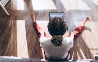 Person sitting on the floor with legs stretched out, working on a laptop in a bright room