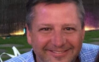 Smiling man outdoors in the evening, wearing a blue checkered shirt.
