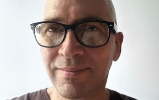 Close-up portrait of a smiling person wearing black glasses and a dark t-shirt in front of a plain light background.