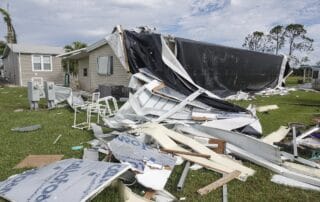 Mobile home severely damaged with debris scattered across a grassy yard after a storm.