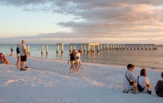 People relaxing and walking on a sandy beach at sunset near a long concrete pier over calm ocean water.