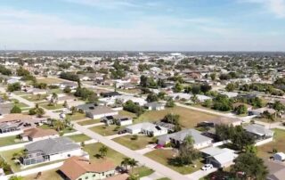 Aerial view of a suburban neighborhood with single‑family homes, green lawns, trees, and straight residential streets under a clear sky.