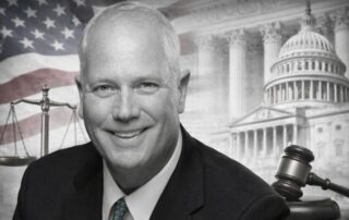 Smiling middle‑aged man in a suit in front of a collage of the u. S. Flag, government buildings, the scales of justice, and a judge’s gavel.