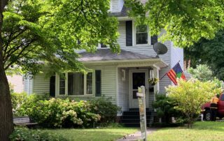 Two-story white house with front porch, lush green yard, and real estate for-sale sign in the front lawn.