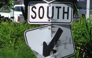 Damaged southbound road sign with a bent directional arrow in front of roadside greenery