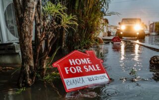 Home for sale sign partially submerged in a flooded street as a vehicle approaches with headlights on.