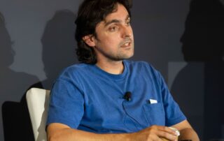 Man in a blue t‑shirt speaking while seated, wearing a lapel microphone.